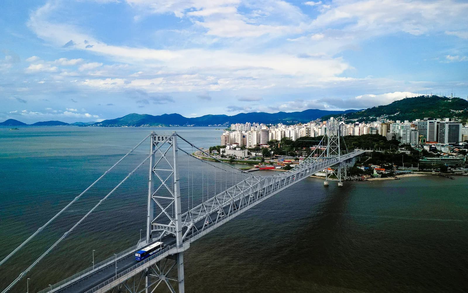 Vista aérea da Ponte Hercílio Luz, com um ônibus na pista e o centro de Florianópolis ao fundo